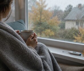 Person mit Decke und Teetasse am Fenster, krank zu Hause