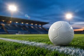 Weißer Fußball auf der Seitenlinie eines Rasenplatzes, dahinter ein leeres Stadion mit Flutlicht