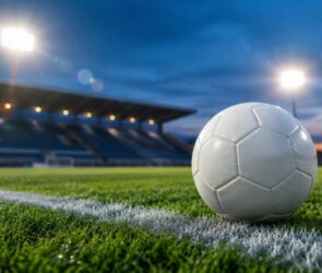 Weißer Fußball auf der Seitenlinie eines Rasenplatzes, dahinter ein leeres Stadion mit Flutlicht