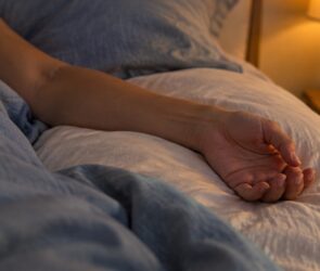 Unterarm einer Person liegt entspannt auf einem weißen Kissen im Bett, leicht gebeugt, im warmen Licht einer Nachttischlampe